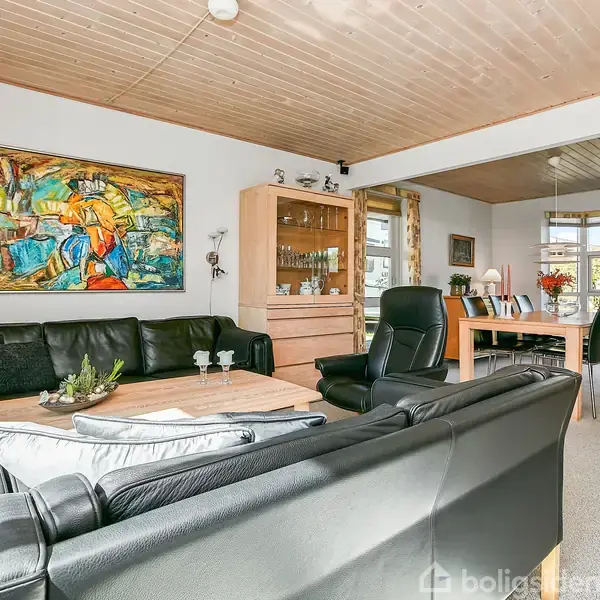 Sort læderhjørnesofa grænser op til en lys stue, hvor lyse trælofter, farverig kunst på væggen og et spisebord med stole ses i baggrunden.