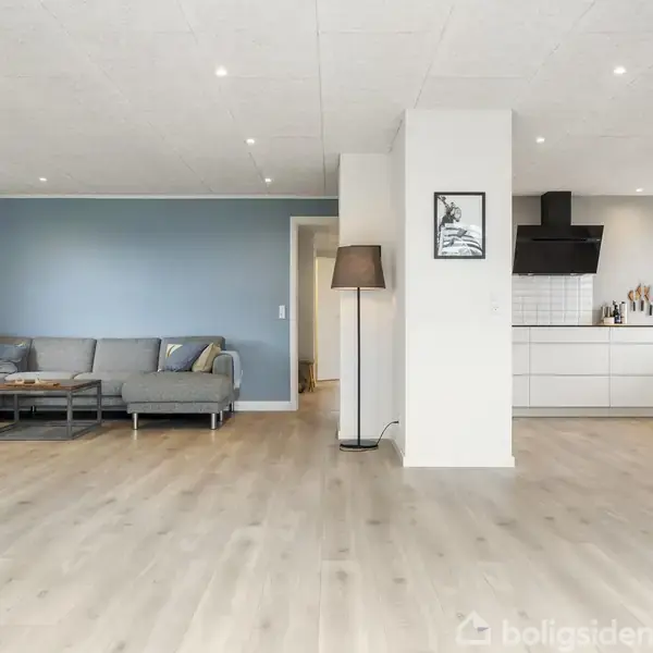En moderne stue med en grå hjørnesofa placeret ved en blå væg. Køkkenet, med hvide skabe, ligger i forlængelse. Et spisebord med stole ses i forgrunden.