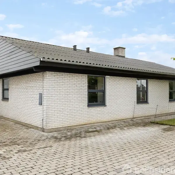 Et enetages hus med hvid murstensfacade står på en flisebelagt indkørsel. Der er græsplæne til højre, og en blå himmel med få skyer danner baggrund.