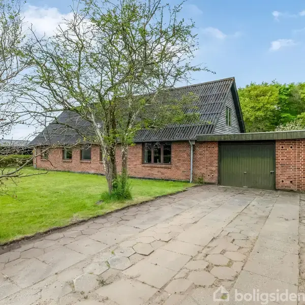 Et rødt murstenshus med en grøn garageport stående stille ved siden af en flisebelagt indkørsel. Omgivet af græsplæne og træer under en blå himmel med spredte skyer.