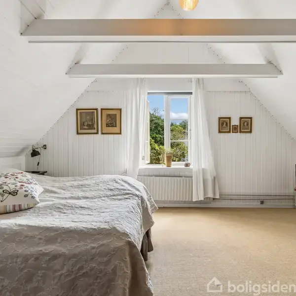 Et lyst soveværelse med skrå vægge har en stor seng til venstre, en hylde med bøger til højre, og et centralt placeret vindue med udsigt til træer og himmel.