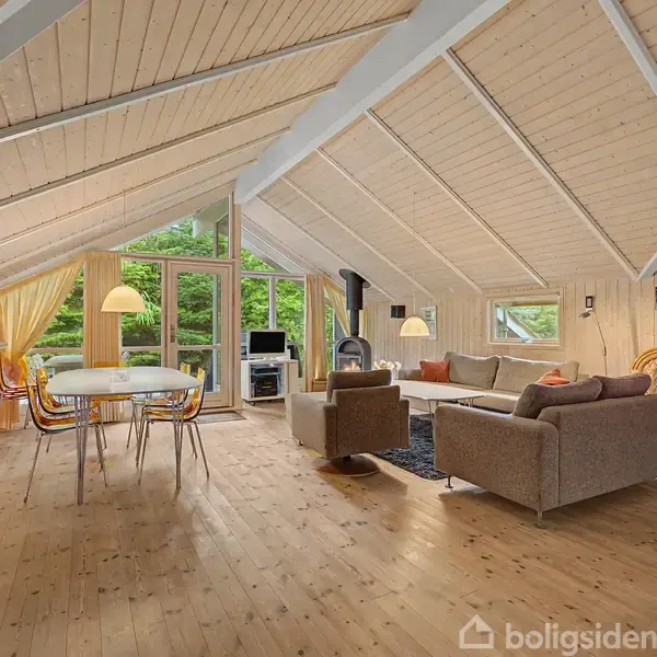 Stue med spisebord og stole midt i rummet, omgivet af trævægge og loft. Store vinduer med udsigt til grønne træer. Komfortable sofaer og pejs i hjørnet.