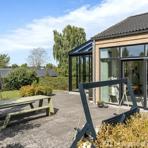 Terrasse med træbord og parasol, omgivet af have, ved siden af moderne hus med glasfacade. I baggrunden er der træer og himmel.