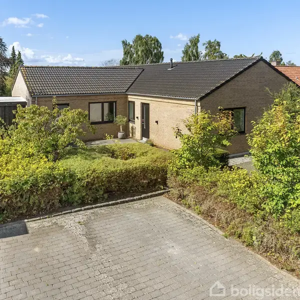En single-story murstenshus omgivet af en velplejet have står i et boligområde. Foran huset er en flisebelagt indkørsel, og der er en garage ved siden af.