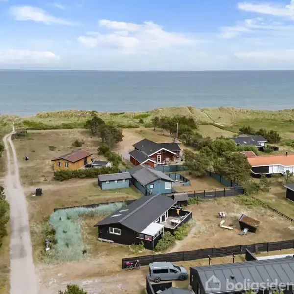 Flere sommerhuse ligger spredt langs en grussti ved kysten. Havet strækker sig i horisonten under en klar blå himmel, omgivet af marker og klitter.