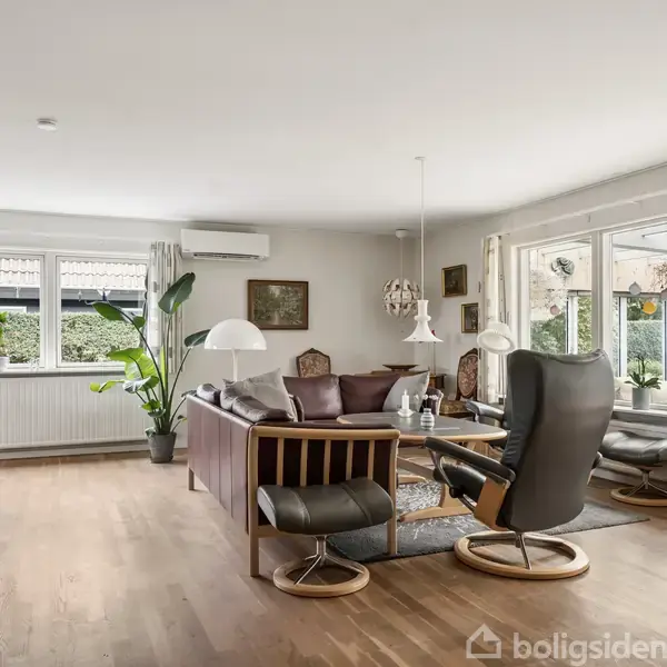 Stue med komfortable stole og sofa, placeret omkring et sofabord. Store vinduer giver udsigt til en terrasse og have. Trægulv og indretning skaber en lys, moderne atmosfære.