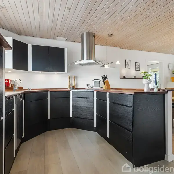 Køkken med sorte skabe og en træbordplade, der danner en vinkel. Emhætte ovenfor midten. I baggrunden spisebord med stole og hvid væg med dekorationer og billeder.