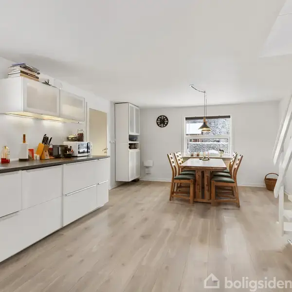 Et lyst køkken med hvide skabe og apparater, hvor ingen synlig aktivitet foregår. Ved siden af står et spisebord med stole i et rum med trægulv og store vinduer.