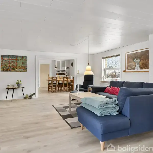 Blå sofa med puder står i en stue, omgivet af et lys træbord, fladskærms-TV, og kunst på væggene. Åben dør fører til et køkken-set-up i baggrunden.