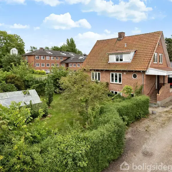 Et rødt murstenshus med rødt tegltag står omgivet af grønne buske og et drivhus. I baggrunden ses flere bygninger, og himlen er let skyet.