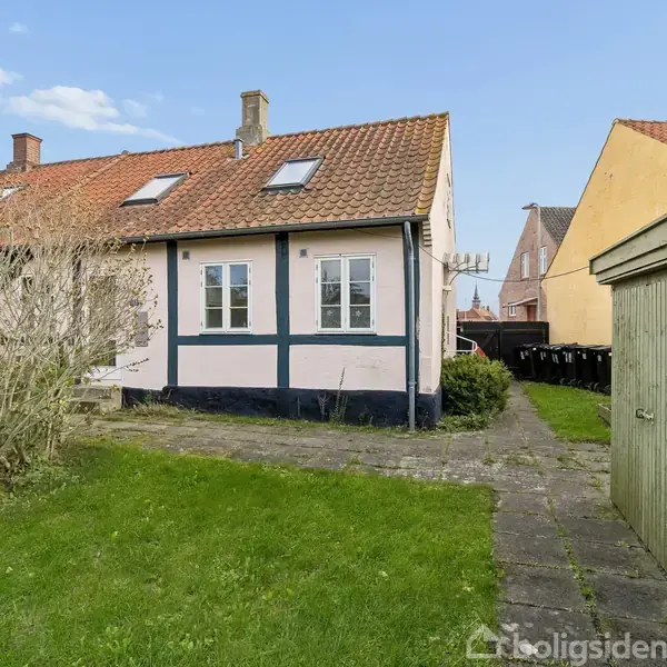 Et bindingsværkshus med rødt tegltag står i en have med græsplæne og flisebelægning. Til højre ses en grøn garagebygning. Himmel er blå med enkelte skyer.