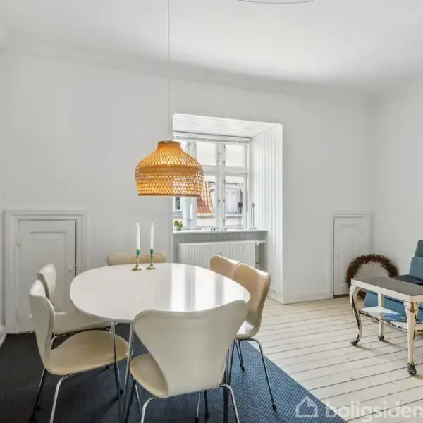 Et rundt spisebord med seks stole står under en hængende lampe, omgivet af en sofa og et lille bord i en lys stue med stort vindue.