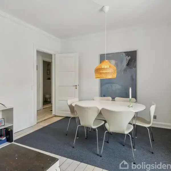 Et rundt spisebord med seks stole står på et blåt tæppe i et lyst rum. En hængelampe i flettet stil lyser over bordet. En reol med bøger står til venstre.