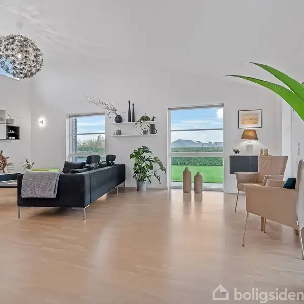 Et moderne stueområde indeholder en sort sofa, grønne planter og loftslampe, omkranset af lyse træmøbler og store vinduer, der giver udsigt til en frodig, grøn have udenfor.