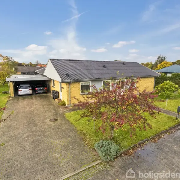 Et enfamiliehus i én etage med gult murværk har sort tag og opkørsel. Der er en carport til venstre med en parkeret bil. Have med træer omgiver huset.