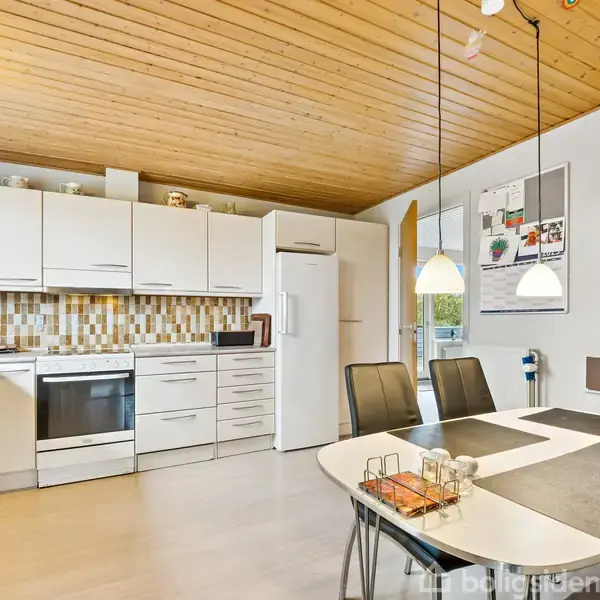 Et lyst køkken med hvide skabe og mosaikfliser, indeholder komfur og køleskab. Et spisebord med sorte stole står midt i rummet. To hængelamper hænger over bordet.