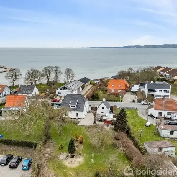 Flere huse med farverige tage ved kysten med udsigt over havet. Parkering i forgrunden med biler. Træer omkring husene skaber en fredfyldt atmosfære.