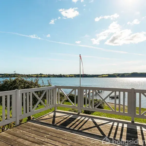 Træterrasse med hvidt rækværk, stille med udsigt over roligt vand og landskab i baggrunden under en klar blå himmel.