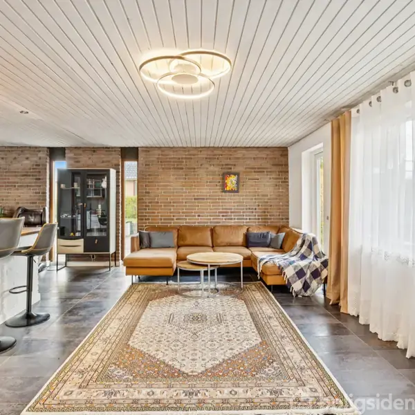 En lædersofa står i en stue med et tæppe på gulvet. En moderne loftlampe lyser rummet, som har murstensvægge og store gardiner ved vinduet.