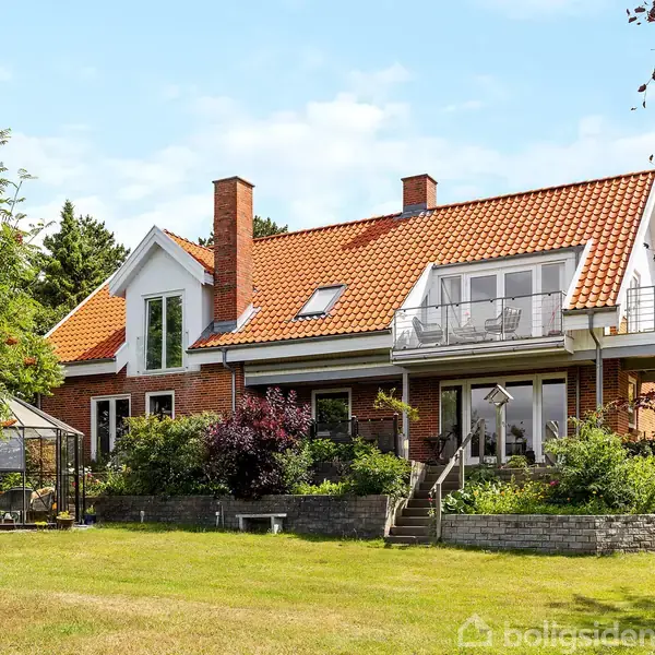 Et rødt murstenshus med rødt tegltag har en balkon og drivhus. Det er omgivet af en velplejet have med græsplæne, buske og træer. Himmel er delvist overskyet.