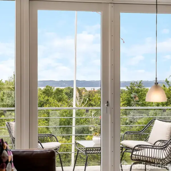 Terrassedøre åbner ud mod en balkon med flettede stole og et bord. Udsigt over grønne træer og en fjord i baggrunden, set fra en stue med sofahjørne.