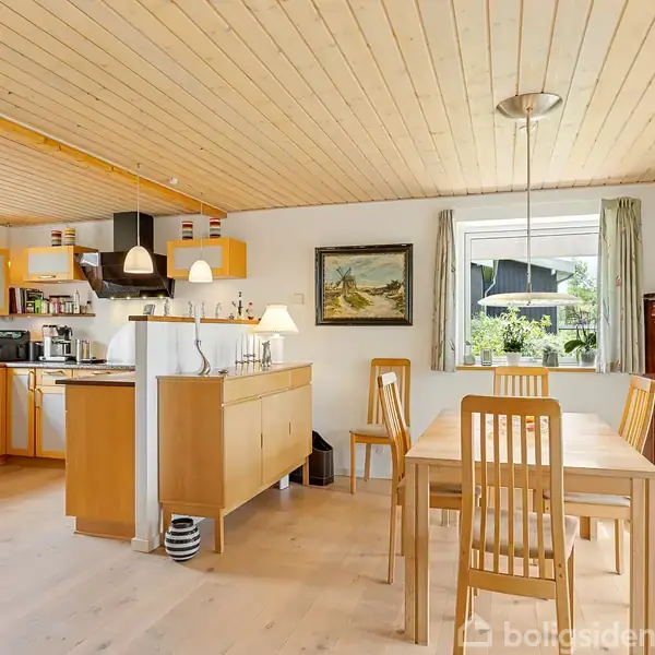 Spisebord og stole står i en lys stue-køkken-alrum med trægulve og træloft. Et skab og et maleri hænger på væggen, mens vinduerne giver udsigt til haven.