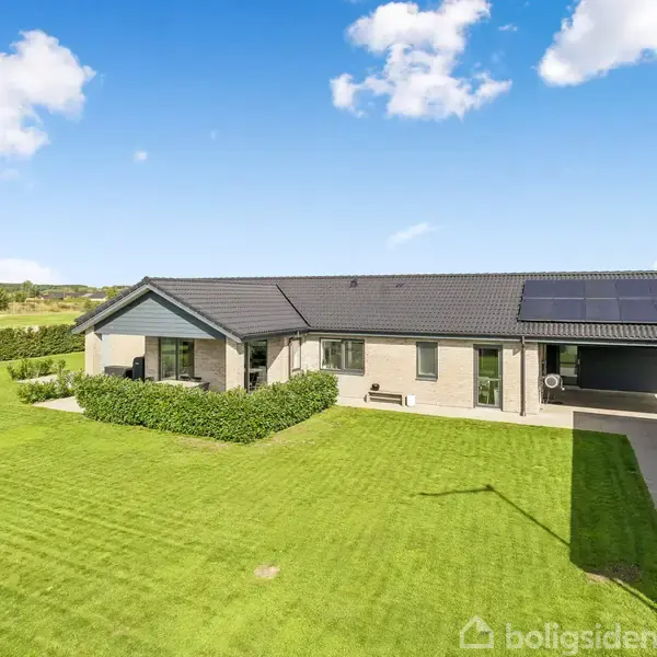 Hus står stille i en solrig have med velplejet græsplæne, omkranset af hække. Solar paneler er installeret på taget. Himlen er blå med få hvide skyer.