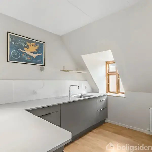 Et moderne køkken med grå skabe, en hvid bordplade og en vask ved et skråtstillet vindue. En plakat forestiller en figur på cykel. Rummet har lyst trægulv.