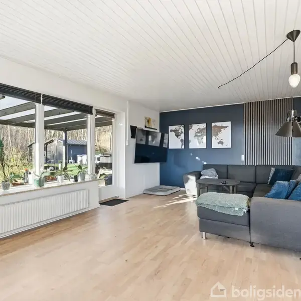 En enkeltfamiliehus med skråt tag ligger omgivet af en terrasse og planter. En bil holder parkeret i en carport til venstre. Et skilt foran huset viser "Til salg".