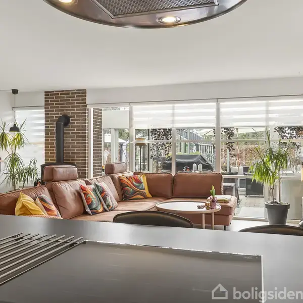 En brun lædersofa med farverige puder står i en moderne stue. Sofaen støder op til store vinduer, omringet af grønne planter og et spisebord i baggrunden.
