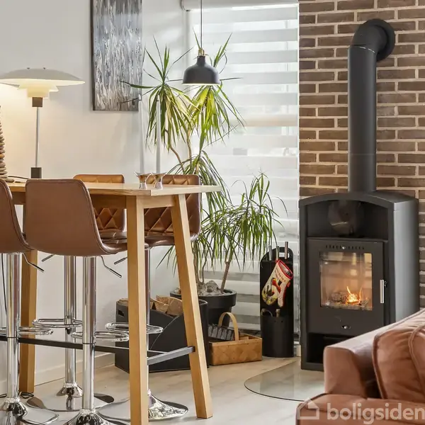 En pejs brænder i hjørnet af en moderne stue med murstensvægge. Et bord med høje stole står ved siden af, omgivet af planter og en sofa.