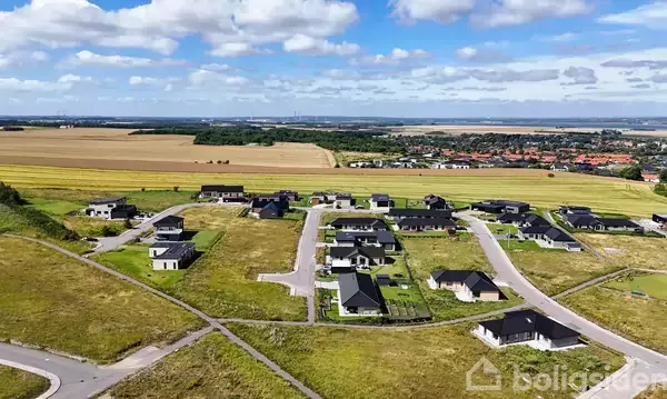 Landskabsbillede viser et boligområde med moderne huse, spredt langs snoede veje. Omgivet af marker og en lille by i det fjerne under en blå, delvist skystækket himmel.