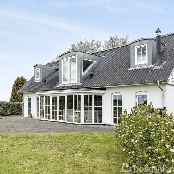 Et hvidt hus med gråt tag og store vinduespartier ser ud mod en grøn have. Træer og buske omgiver huset, der står på en velplejet græsplæne.