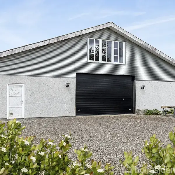 Bygning med grå facader og sort garageport står på grusbelagt areal. Omkranset af grøn vegetation, and et bord-bænkesæt til højre samt en klar blå himmel.