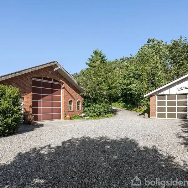 To bygninger står stille på en grusvej, omgivet af grønne træer. Den ene bygning er en stor rødstensgarage med en høj port, den anden en mindre struktur.