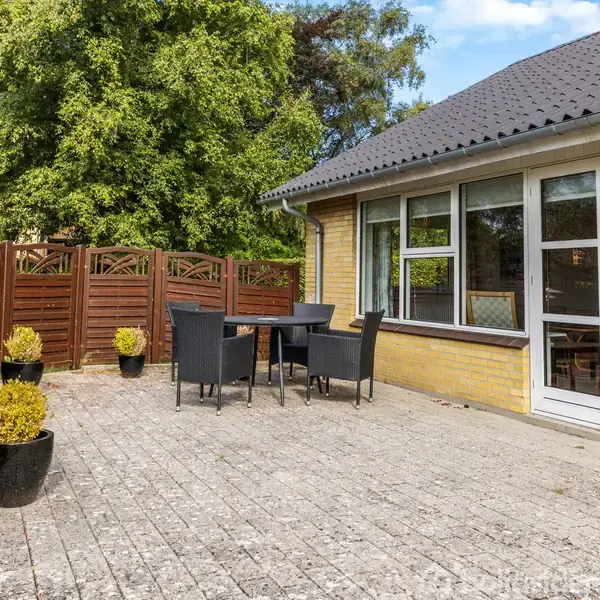 En flisebelagt terrasse med sorte havemøbler, omkranset af gule murstensvægge og træhegn. Flere planter i sorte krukker skaber en hyggelig atmosfære ved siden af et hus.