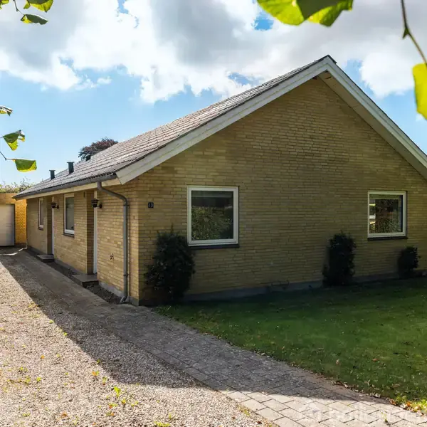 Gulstenshus står stille ved en grusindkørsel, omkranset af grønne buske og træer under en klar blå himmel med skyer.