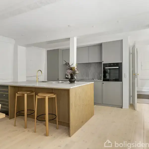 Køkkenø med to træstole står i et moderne køkken med grå skabe og indbygget ovn. En vase med blomster pynter bordpladen. Lyse trægulve og dør åbner til andet rum.