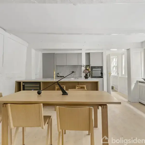 Et lyst køkken med et bord med fire stole i forgrunden. I baggrunden er der et moderne køkken med grå skabe, en ovn og vinduer til højre.