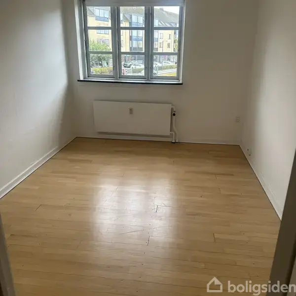 Et tomt værelse med lyse trægulve og hvide vægge. Et vindue viser udsigt til en gade. En radiator er placeret under vinduet. Teksten "boligsiden" er nederst.