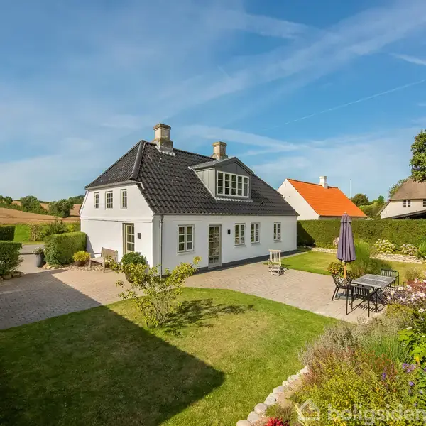 Et hvidt hus med sort tag står i en velplejet have med græsplæne og blomsterbede. Havemøbler med parasol står på en terrasse, og der er blå himmel.