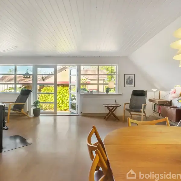 Stue med træmøbler i et lyst rum; spisebord med stole i forgrunden, sofaer ved vindue og skænk ved siden. Store vinduer giver udsigt til en have.