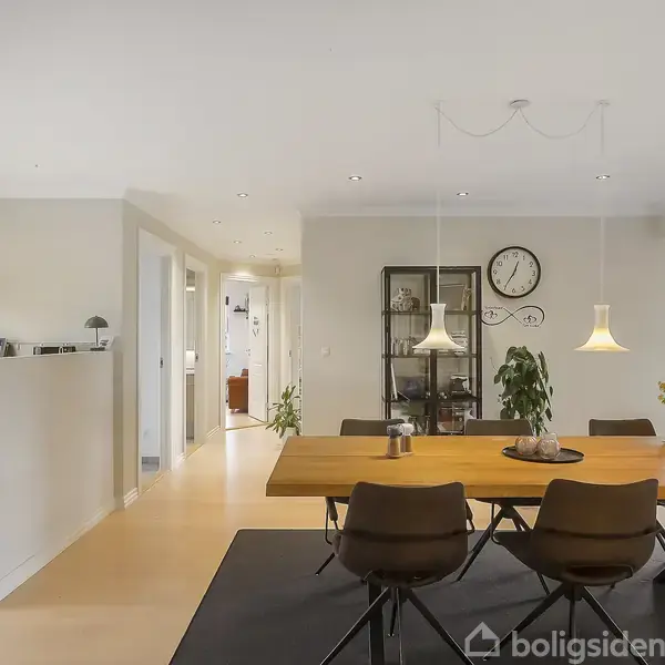 Et spisebord med seks stole står i en spisestue. Lamper hænger over bordet. I baggrunden er der en hylde med planter og ure, og der er en åben forbindelse til køkkenet.
