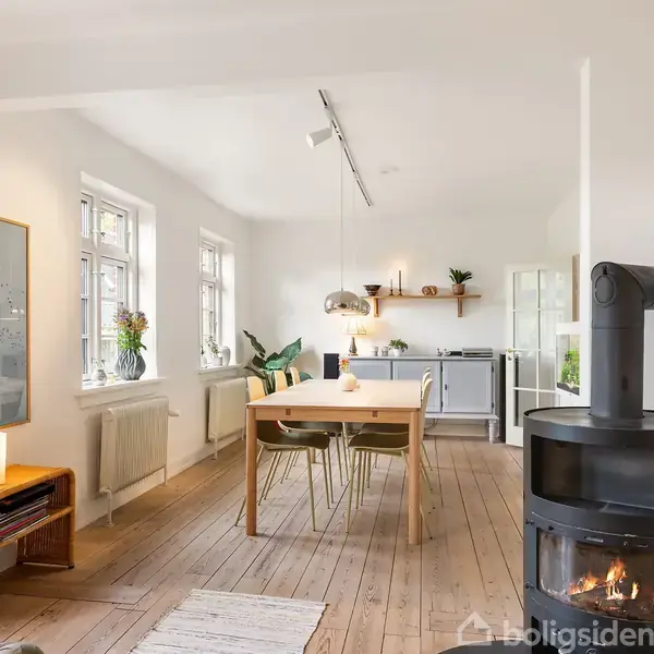 En hyggelig stue med en brændeovn til højre, træbord og stole centralt placeret, omgivet af lyse vægge, store vinduer og grønne planter, giver et moderne, hjemligt indtryk.