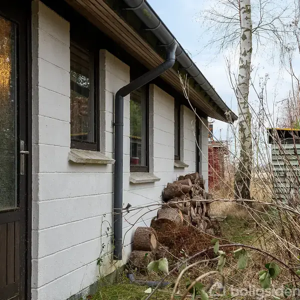 Et hvidt murstenshus står stille ved en have. Ved siden af huset ligger en bunke brændestykker. Et træ og buske omkranser området.
