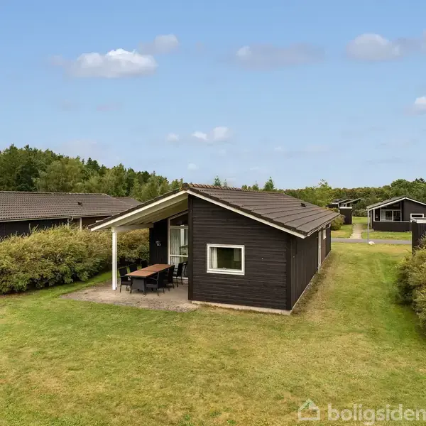 Et sort træhus med et lille overdækket område og havemøbler står på en grøn græsplæne, omgivet af buske og flere lignende huse i det fjerne.