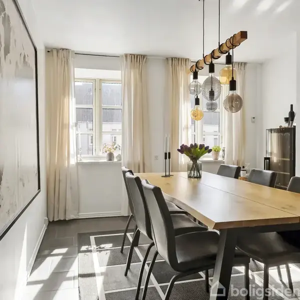 Spisebord i træ er omgivet af seks stole, dekoreret med en vase blomster. Rummet er lyst med hvide gardiner ved vinduet og moderne pendellamper. Kunst pryder væggen.