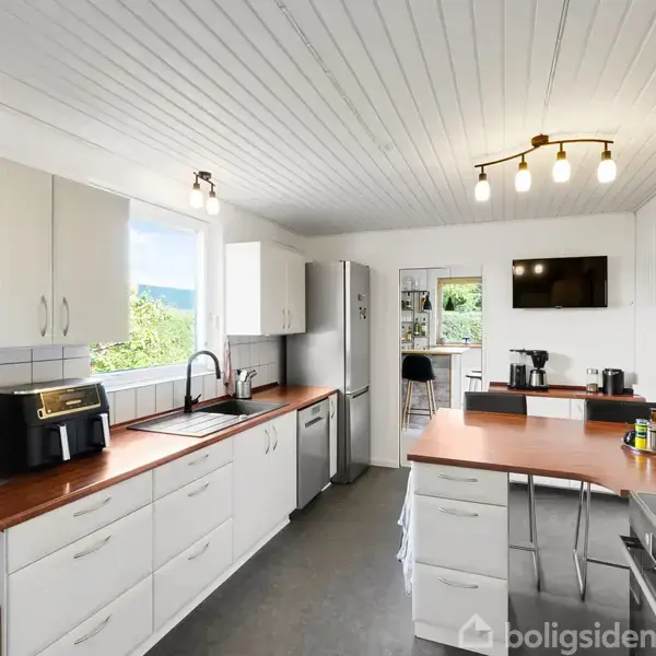 Køkken med hvide skabe og trælignende bordplader. Der står en brødrister og mikroovn på bordet. Et vindue giver udsigt udenfor. En dør fører til en spisestue.