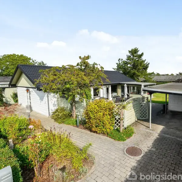 Et enetages hus med hvid facade og mørkt tag ligger i en have med blomsterbede og buske. Der er en carport til højre for huset.
