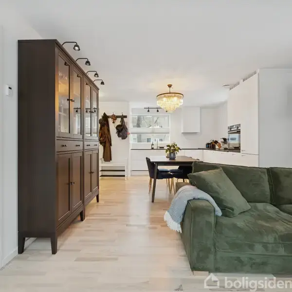 En grøn sofa står i en stue med en åben planløsning mod et lyst køkken. En stor lysekrone hænger over spisebordet, og der er et mørkt skab langs væggen.